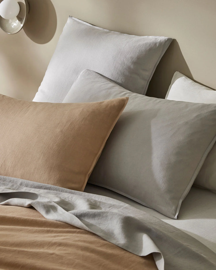 Neutral layered bedding setup featuring linen pillowcases and flat sheet in soft biscuit and silver tones with textured weave, minimalist bedroom styling with warm natural light