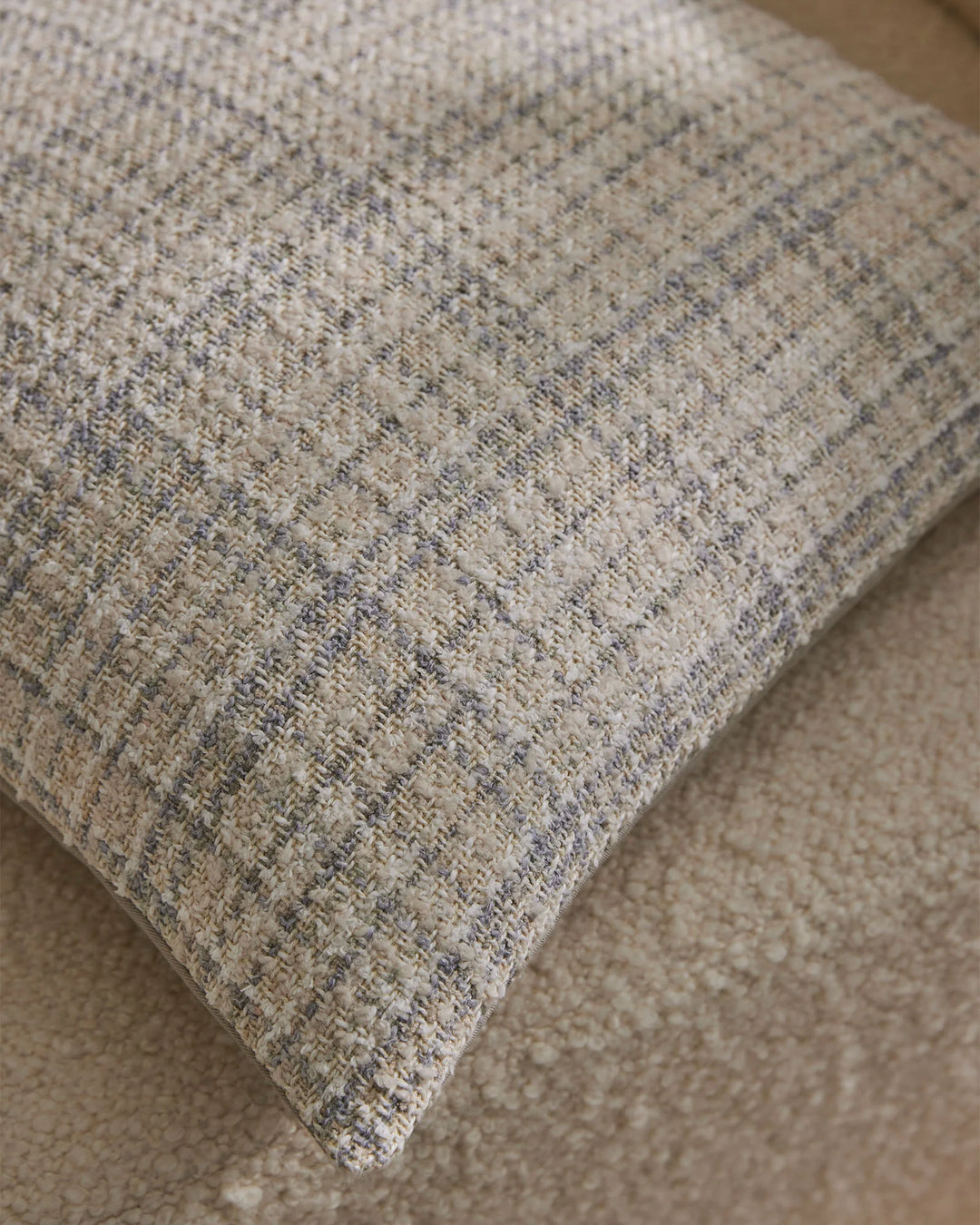 Close-up of neutral textured cushion with herringbone weave pattern in soft greens and greys, layered on warm sand-toned upholstery