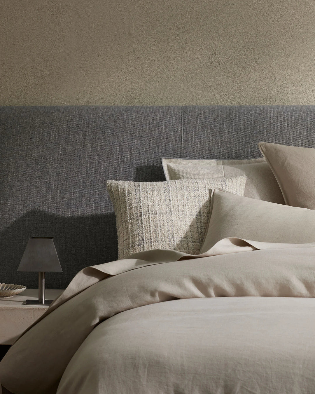 Neutral bedroom styling with cream linen bedding, grey upholstered headboard, and layered cushions in natural textures including a chunky knit pillow