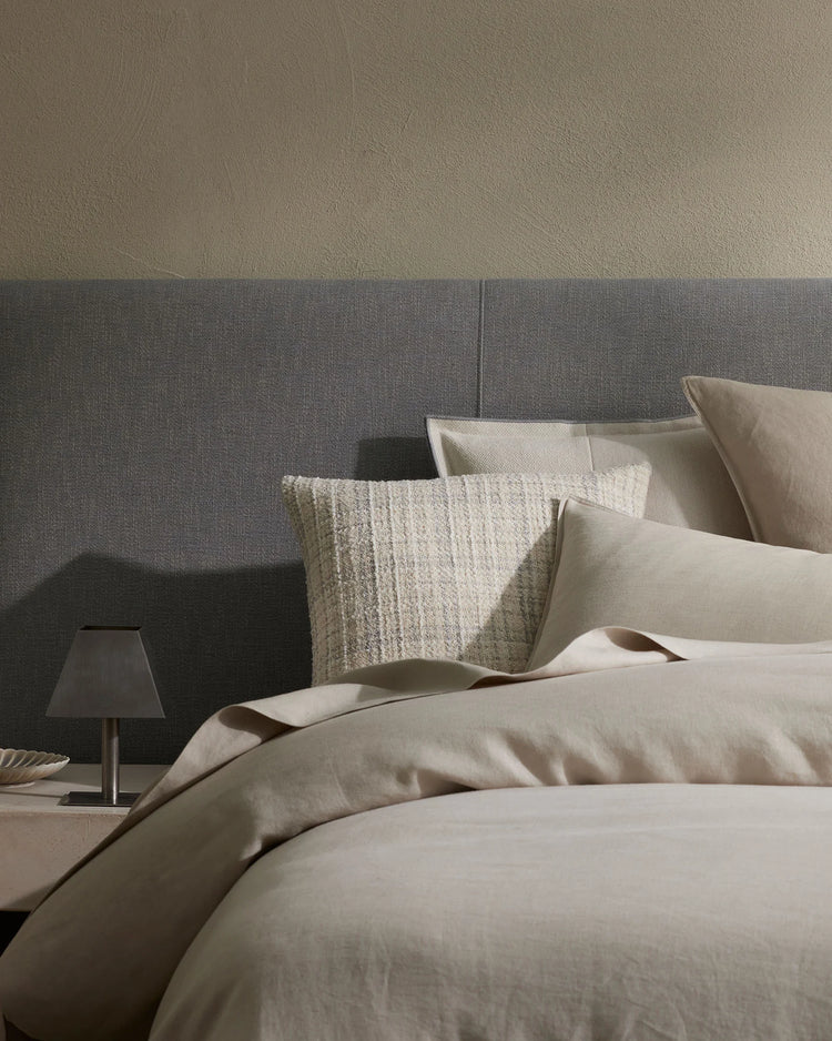 Neutral bedroom styling with cream linen bedding, grey upholstered headboard, and layered cushions in natural textures including a chunky knit pillow