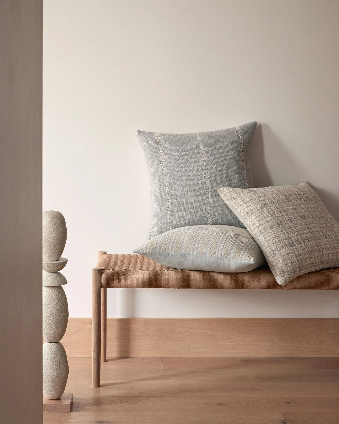 Neutral minimalist bedroom scene with layered linen cushions in soft grey, cream and natural tones on wooden bench, complemented by textured wall art