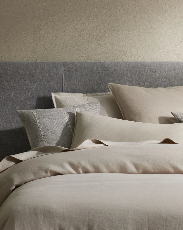 Neutral bedding scene with cream linen sheets and Maddalena cushions layered against a textured grey upholstered headboard, emphasizing soft monochromatic tones and natural textile textures