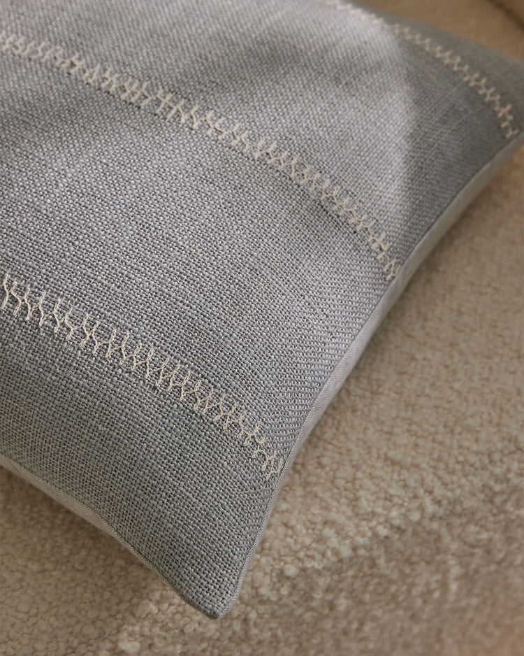 Maddalena Glacier cushion draped on neutral textured carpet, showcasing pale grey linen-polyester blend with delicate whipstitched edging in soft monochromatic styling