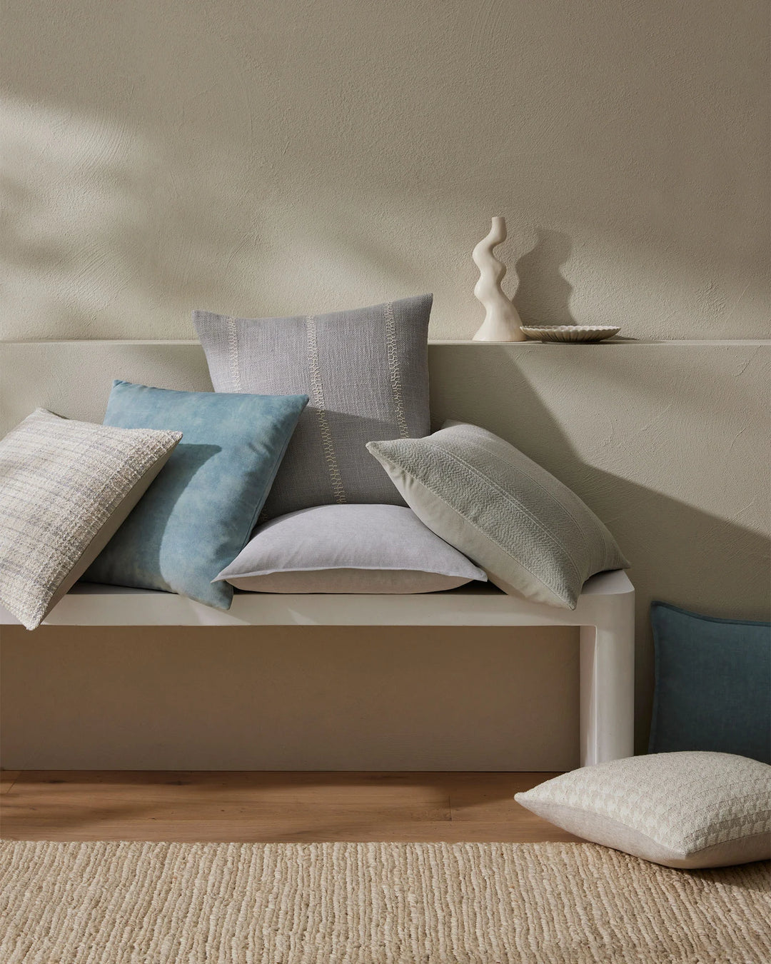 Neutral bedroom styled with layered cushions in cream, grey and soft blue tones on a beige upholstered bed, featuring textured linens and natural jute rug beneath