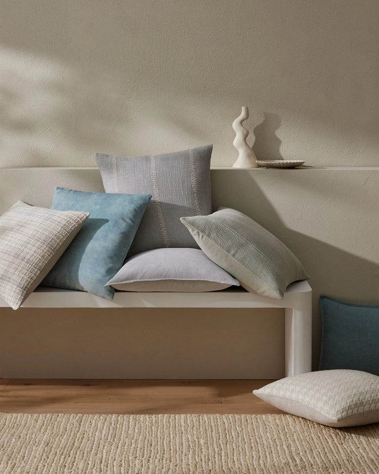 Neutral bedroom styled with layered cushions in cream, grey and soft blue tones on a beige upholstered bed, featuring textured linens and natural jute rug beneath