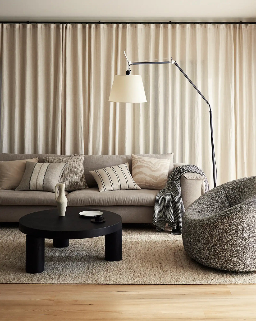 Neutral living room with beige linen curtains, taupe sofa styled with striped and textured cushions, charcoal throw blanket, and natural fibre accents in a minimalist, contemporary aesthetic
