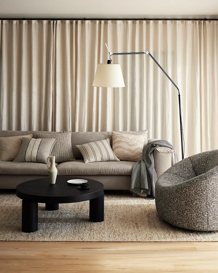 Neutral living room with beige linen curtains, taupe sofa styled with striped and textured cushions, charcoal throw blanket, and natural fibre accents in a minimalist, contemporary aesthetic