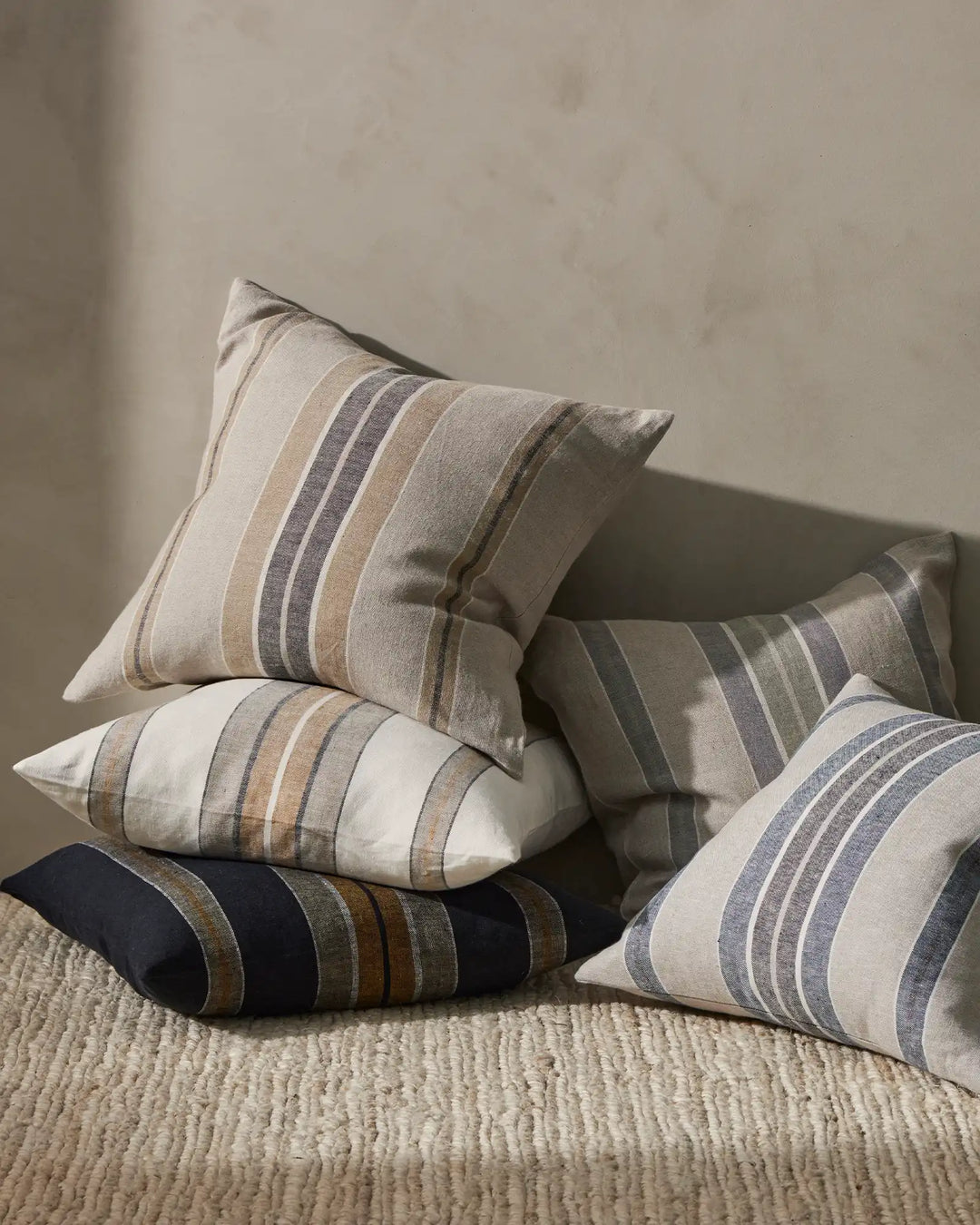 Striped linen cushions in sage, clay, amber, denim and shadow tones arranged on a natural woven texture, minimalist neutral styling with soft directional light