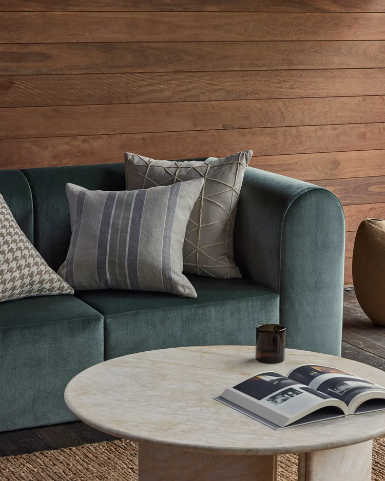 Warm wood-paneled living room with sage-green upholstered sofa styled with striped linen and geometric patterned cushions, marble coffee table, and natural fiber textures