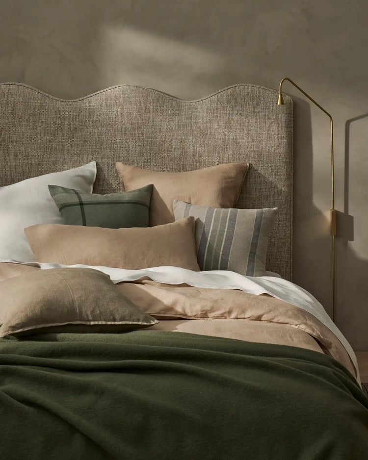 Neutral bedroom layering with sage Franco Cushion and striped Dante cushion against textured linen headboard, warm beige bedding, and forest green throw in muted earthy tones