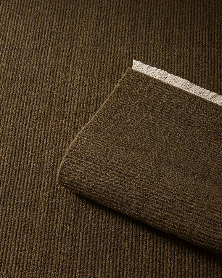 Olive-toned wool rug in ribbed texture, layered with darker Fabien moss-coloured throw showing natural fringing detail against neutral background