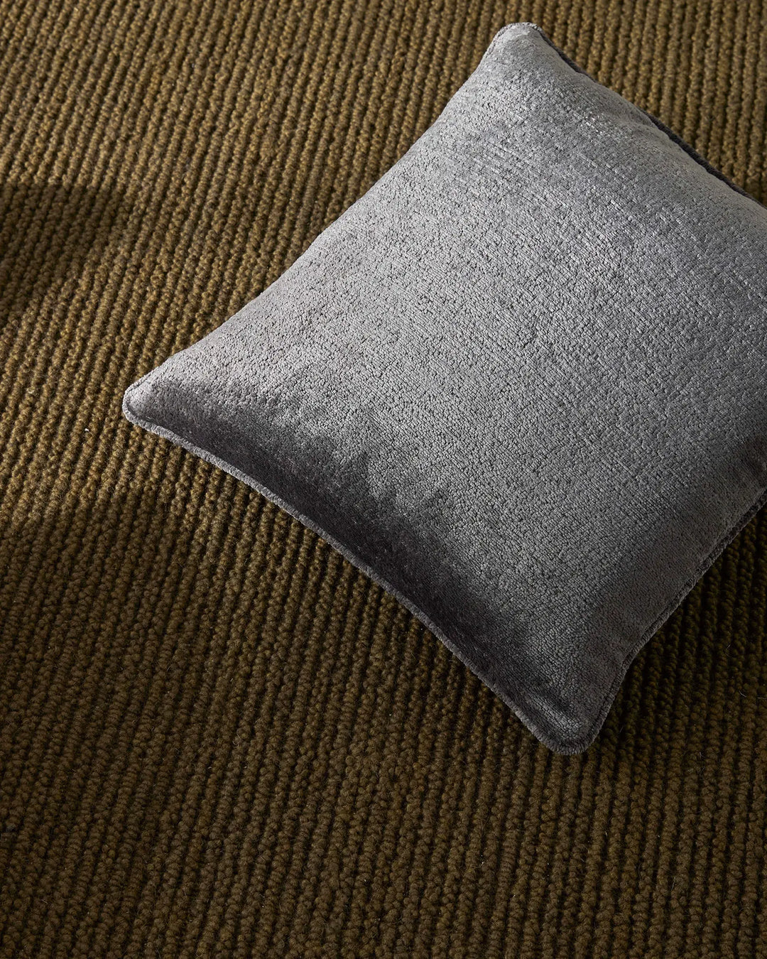 Grey linen cushion resting on earthy moss-toned wool rug with linear weave texture, neutral colour palette interplay