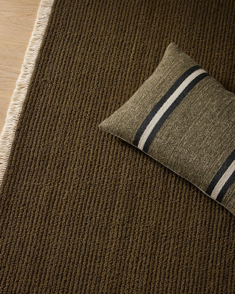 Olive-toned wool rug with ribbed texture and cream fringe, styled with striped cushion in earthy neutrals and charcoal stripes on light wood floor