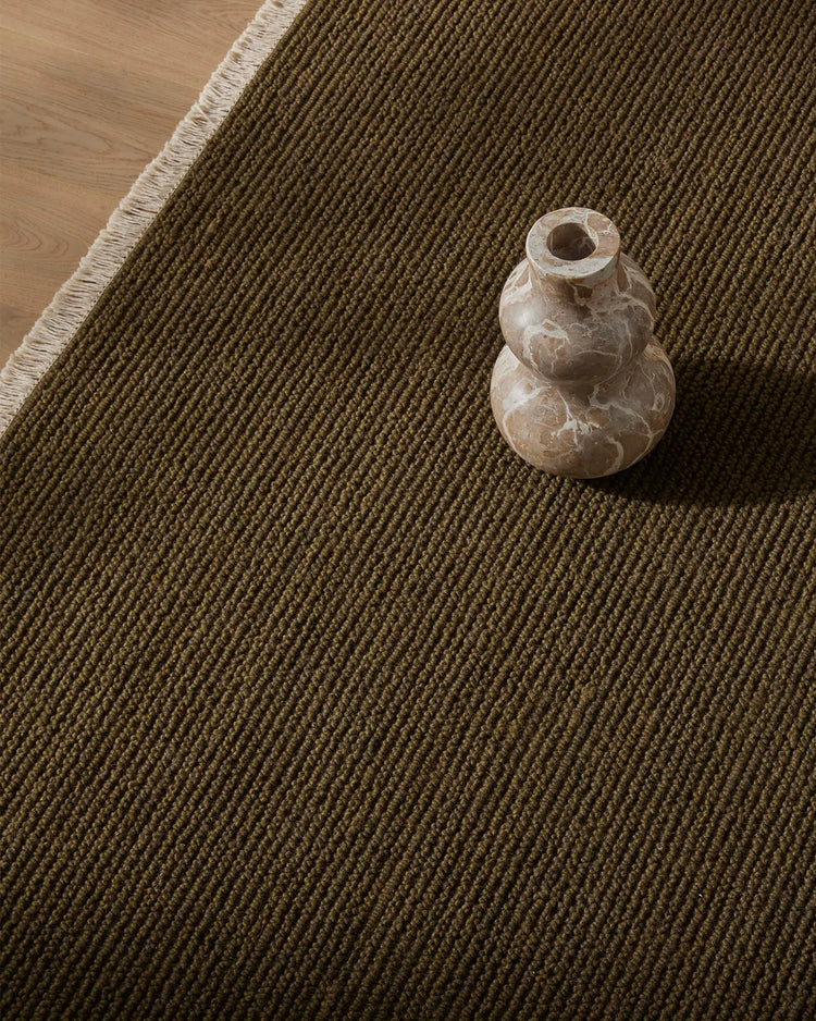 Olive wool rug with tight diagonal weave texture, cream bound edge, paired with warm timber flooring and sculptural stone vase styling