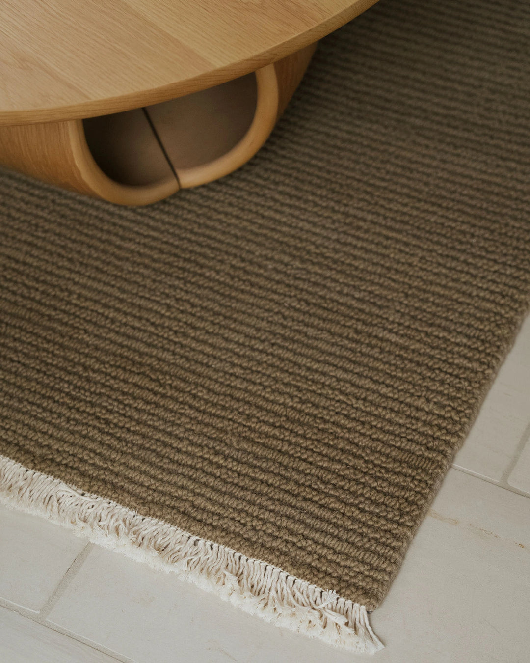 Warm-toned wool area rug with linear texture and cream fringe trim, styled with natural wood furniture in modern minimalist interior