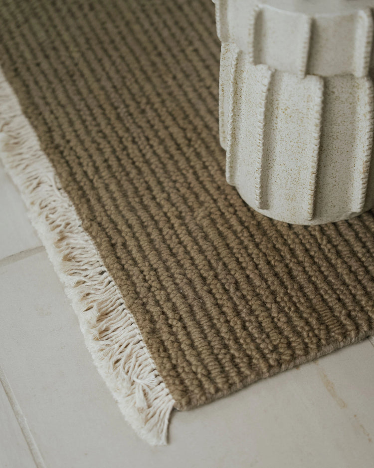 Neutral wool rug with ribbed texture and cream fringe beside white sculptural home accessories on pale tile flooring, earthy minimalist interior styling