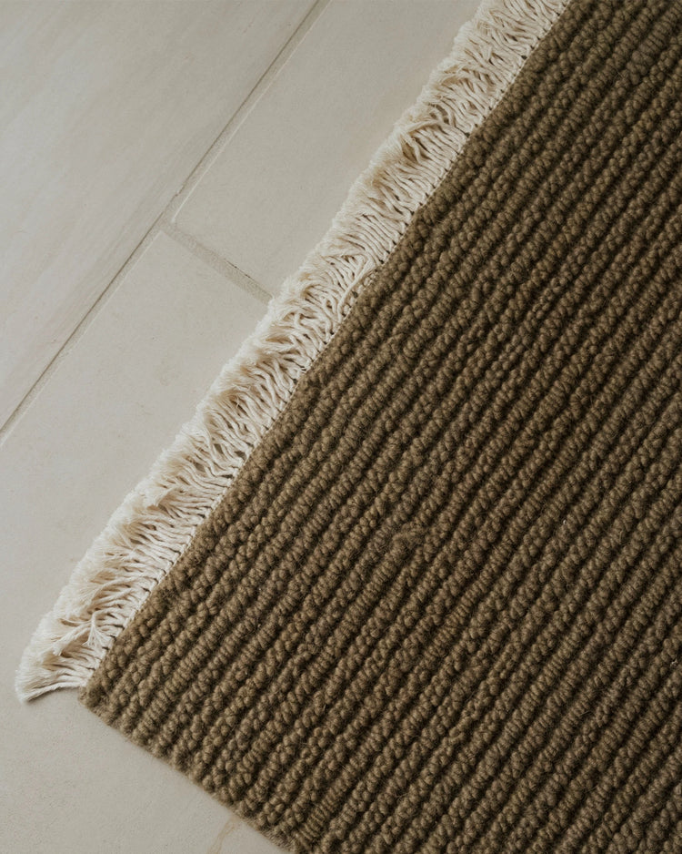 Neutral wool rug with ribbed texture and fringed edge on pale wooden floor, showing earthy taupe and cream tones in minimalist Scandinavian styling