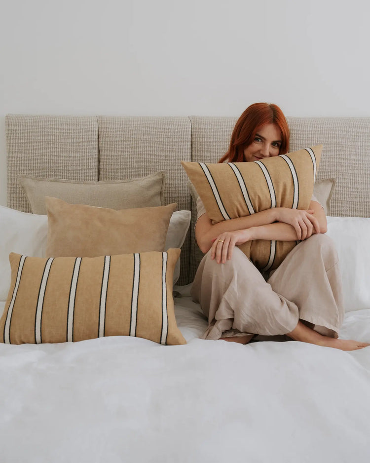 Neutral bedroom styled with warm ochre and charcoal striped cushions layered on natural linen sofa, taupe bedding and white sheets creating a calm, minimalist aesthetic