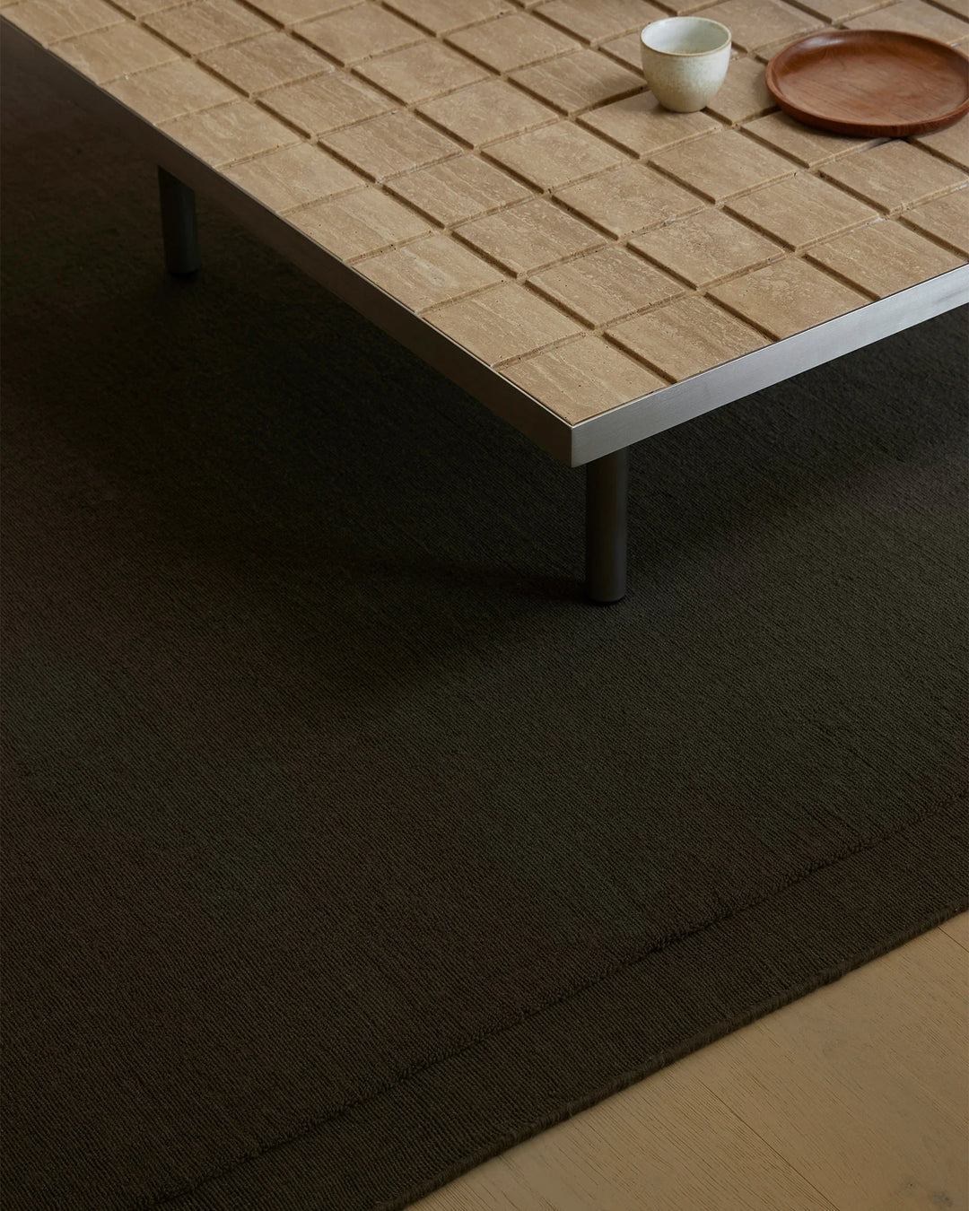 Minimalist dining space with natural wood table, charcoal wool area rug, and neutral ceramics. Warm timber tones contrast with deep charcoal Cassio rug in understated Japandi aesthetic.