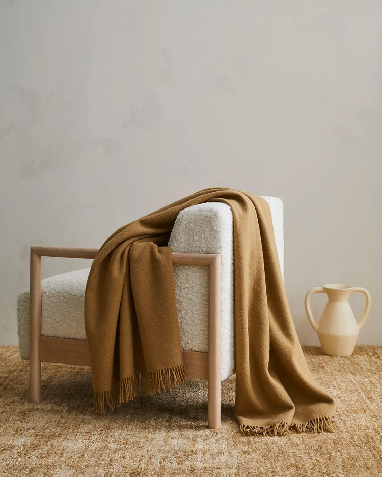Nevis Camel lambswool throw in warm biscuit brown, draped over natural wood chair, tassel-fringed edges visible, by Weave Home