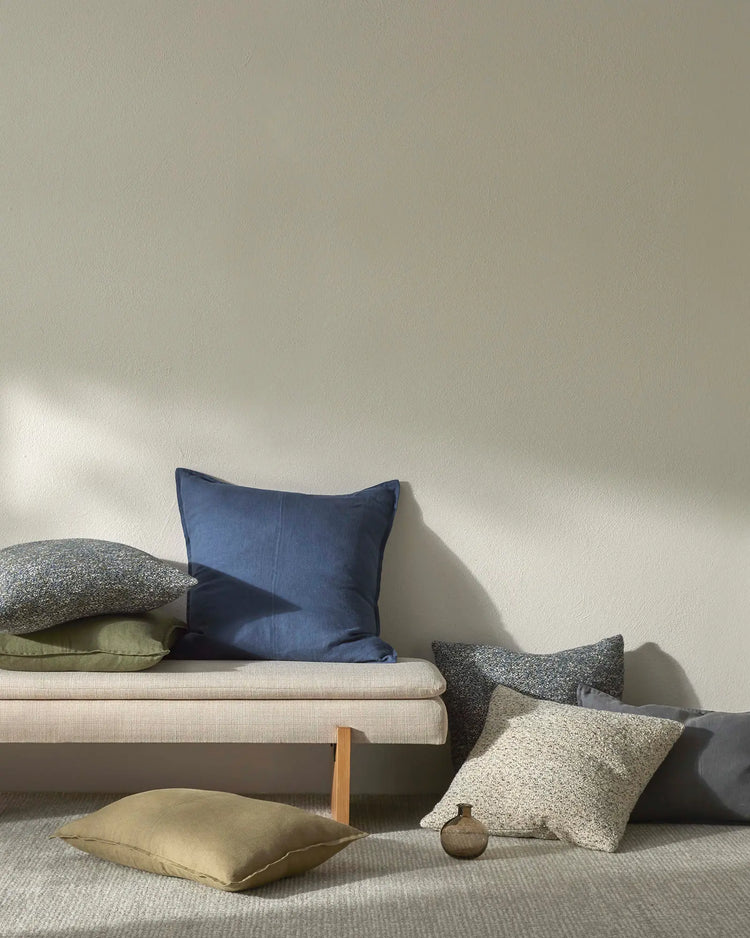 Minimalist living room styled with neutral textiles: cream linen sofa, sage and charcoal linen cushions, ochre silk pillow, and soft grey wool rug against white walls with natural light