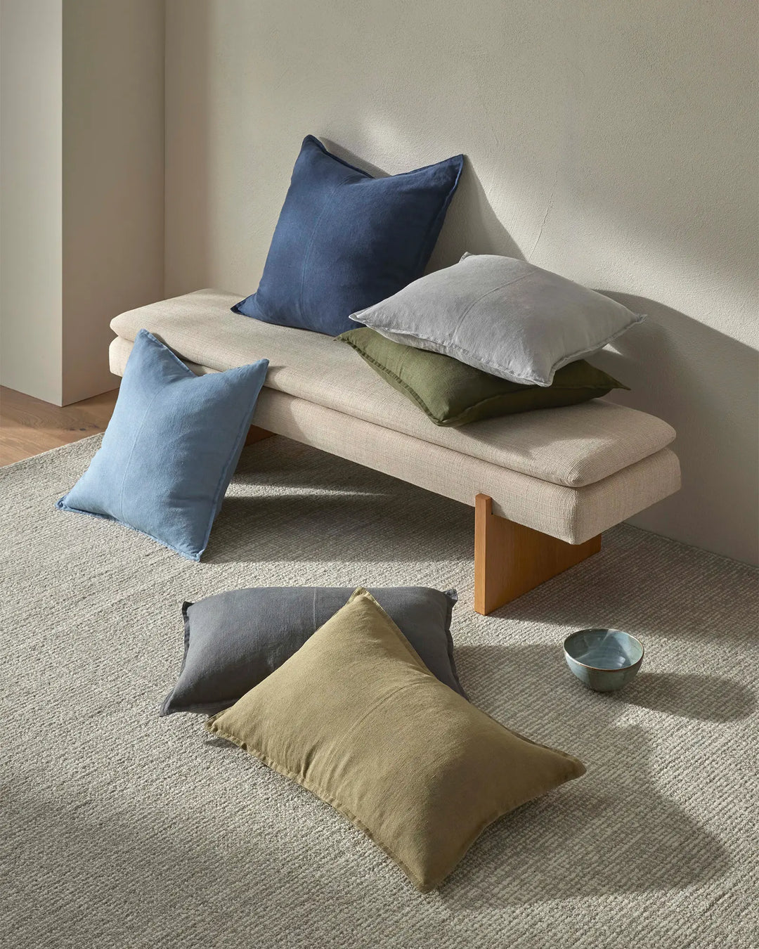 Minimalist living room with cream linen bench and scattered linen cushions in navy, soft blue, sage green, charcoal and warm taupe tones against natural jute flooring and white walls