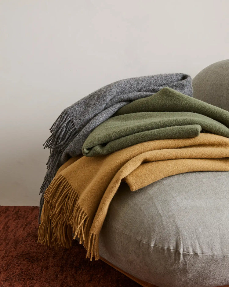 Layered wool throws in charcoal, sage green and camel draped over a linen cushion, earthy neutral palette with rich burgundy rug and soft grey backdrop