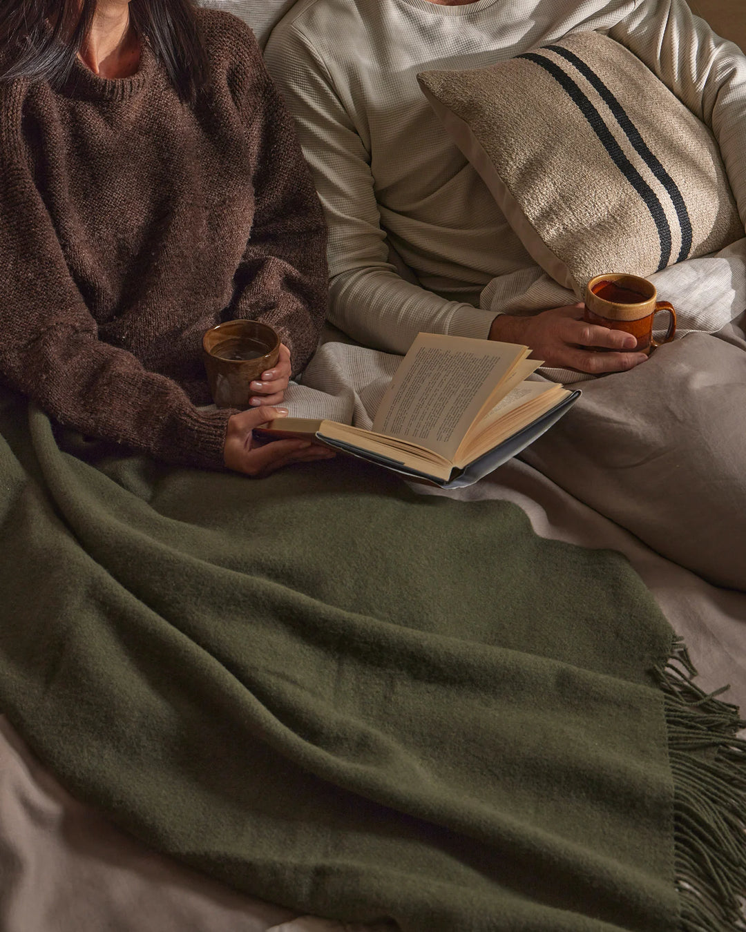 Cosy reading moment with earth-toned textiles: chocolate and sage wool throws layered on neutral bedding, striped linen cushion, warm ceramic mugs, and open book capturing slow-living aesthetics