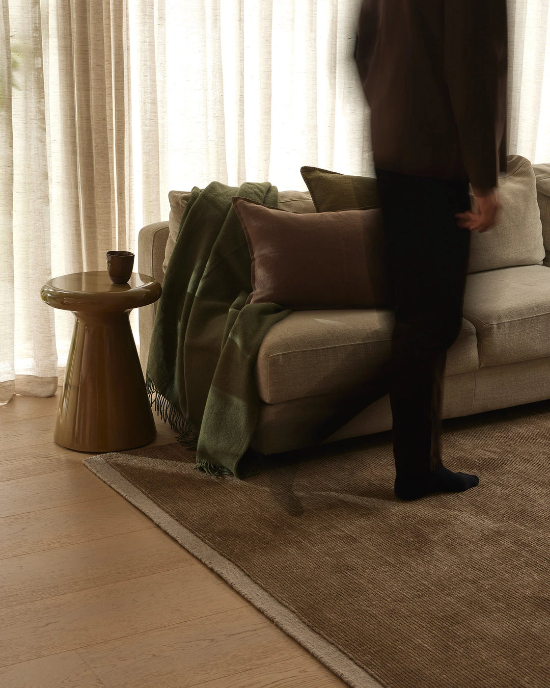 Earthy living room with beige linen sofa, sage-toned wool throw, and layered neutral textures. Warm wood accents, soft natural light through sheer curtains, and natural fiber rug create calm, organic aesthetic.
