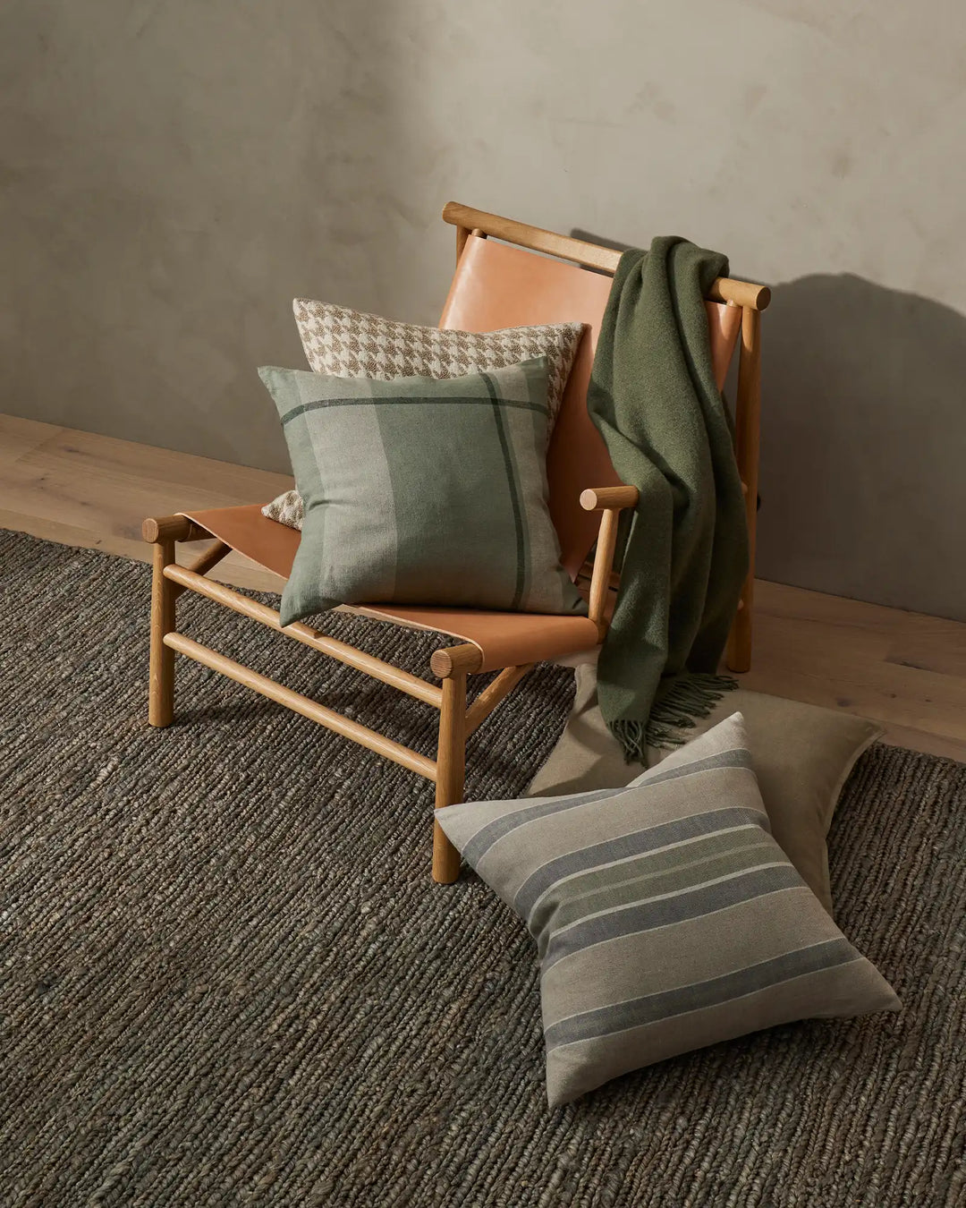 Scandinavian-inspired living space with earthy tones: sage green and warm cognac leather lounge chair layered with striped and houndstooth patterned cushions, textured throw, and neutral striped floor cushion on charcoal wool rug.