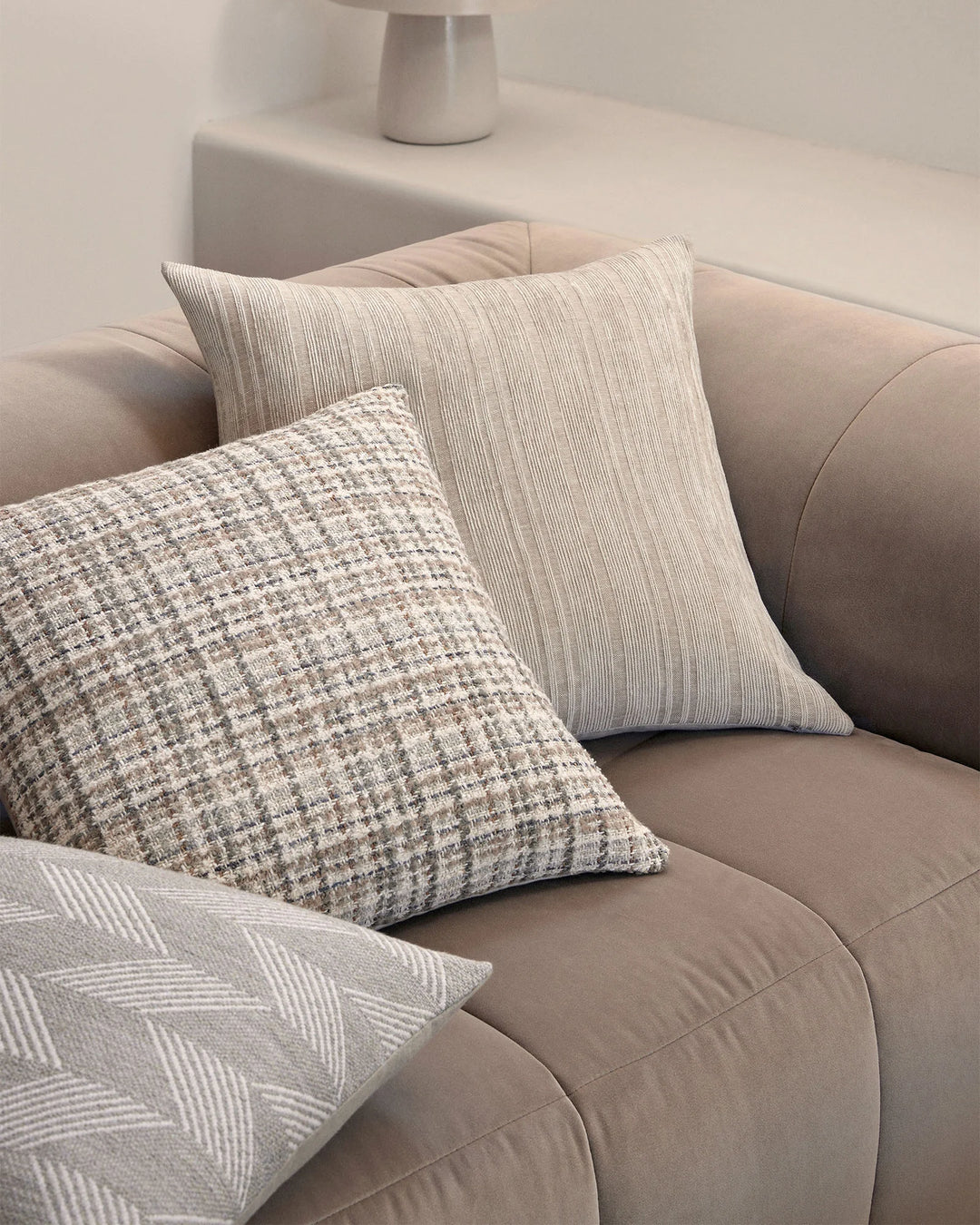 Neutral layered cushion styling on a curved beige sofa featuring textured linens, woven jacquard, and striped patterns in cream, natural, and soft taupe tones