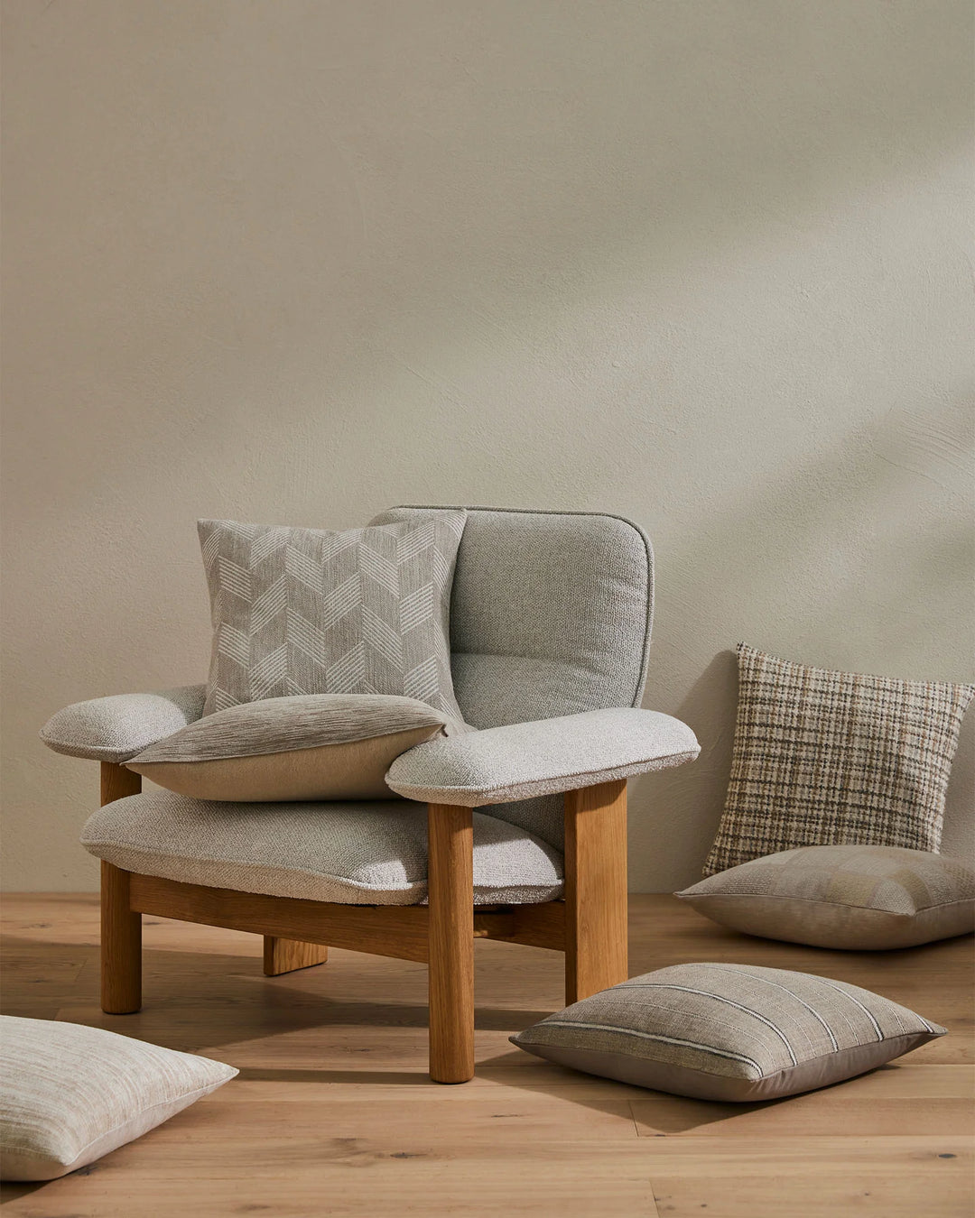 Neutral minimalist interior with wooden-frame chair styled with beige and grey cushions in geometric and textured weaves, displayed on warm timber flooring against soft taupe wall.