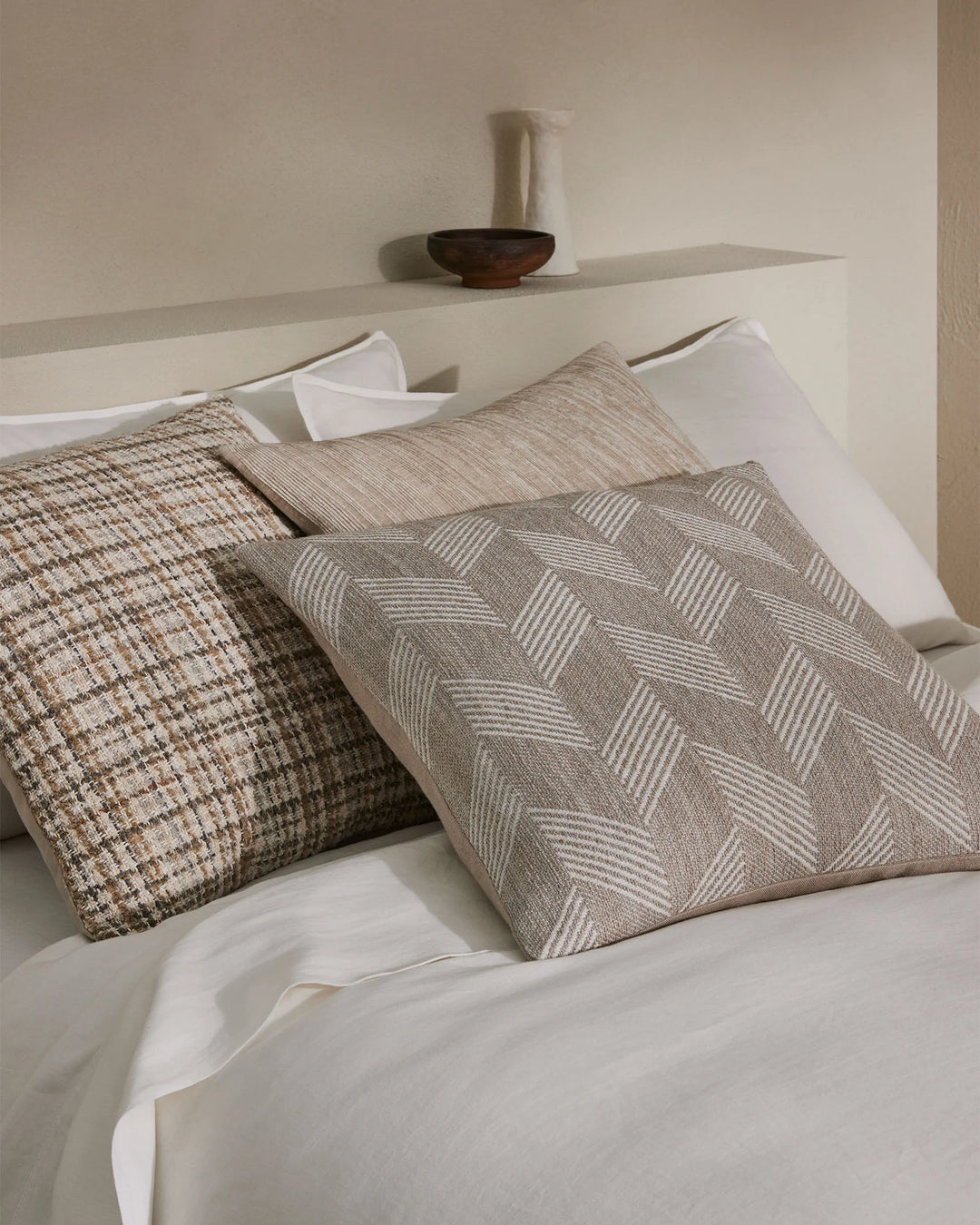 Neutral bedroom styling with layered textured cushions in beige and sage tones on white bedding, featuring geometric weaves and linear patterns alongside a minimalist wooden bowl accent.