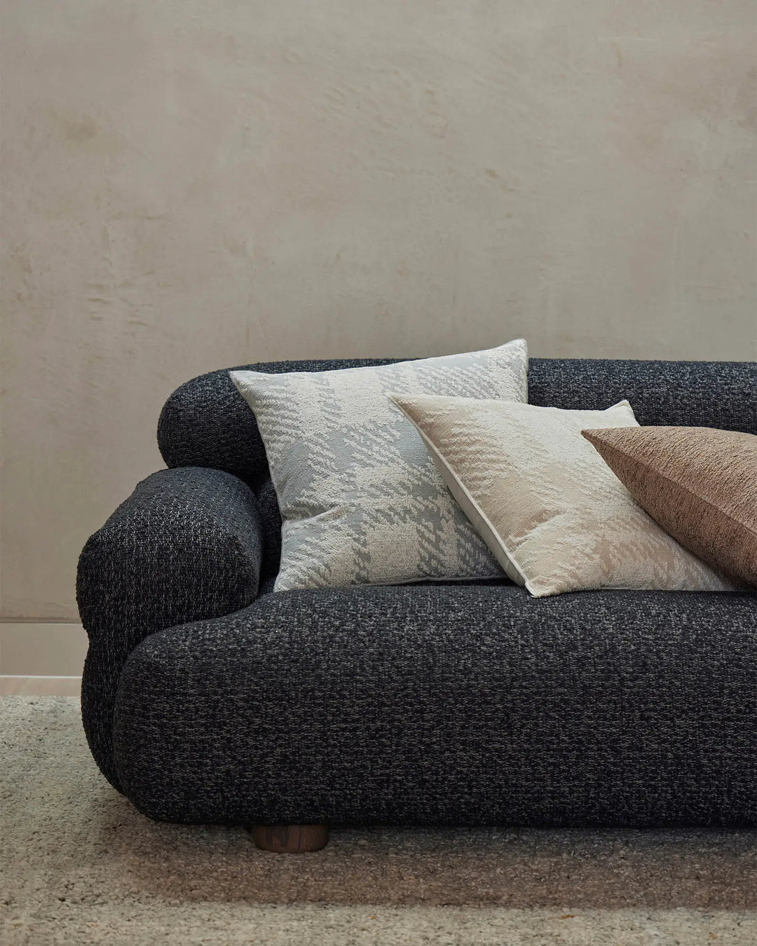 Charcoal curved sofa styled with neutral textured cushions in cream, taupe and warm brown tones, creating a minimal Scandinavian aesthetic with soft layered fabrics