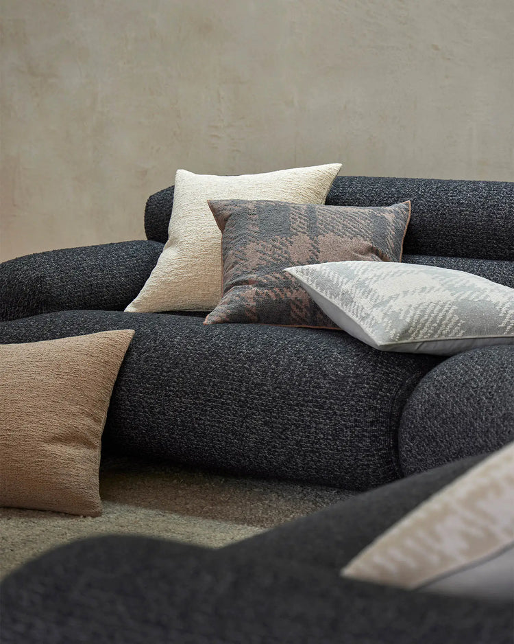 Charcoal curved sofa styled with layered cushions in warm taupe, cream, and dusty blue tones against a textured neutral wall, showcasing woven textile depths and contemporary interior design.