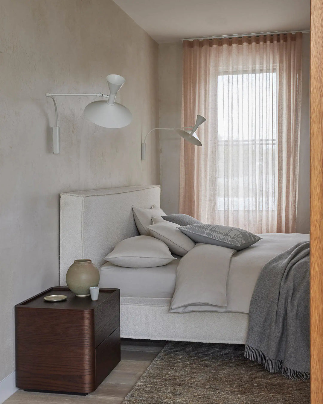 Minimalist bedroom styled with neutral bedding, grey cushions, and layered textures including a jute rug, set against blush curtains and warm wood tones