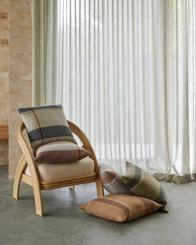 Neutral minimalist bedroom with wooden armchair, striped and solid Dante cushions in terracotta, tobacco and linen tones, sheer floor-to-ceiling curtains, pale limestone wall, concrete flooring