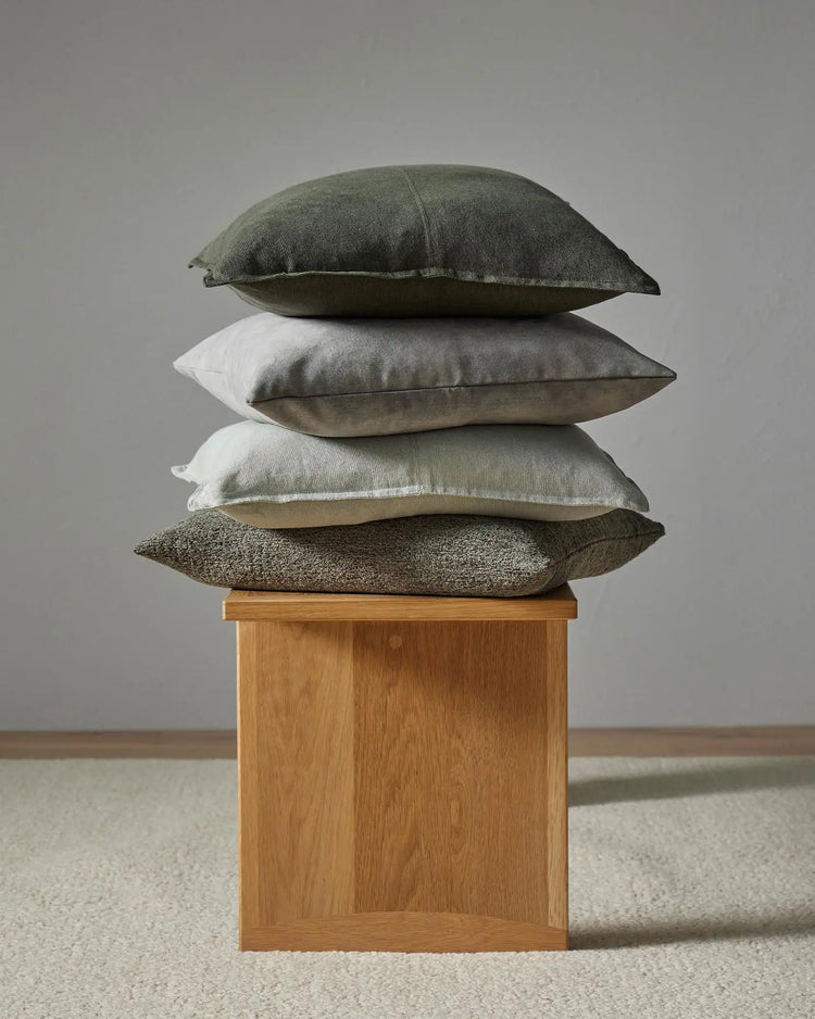 Stacked linen cushions in sage, cream and charcoal tones with textured weaves, displayed on natural timber base against neutral backdrop