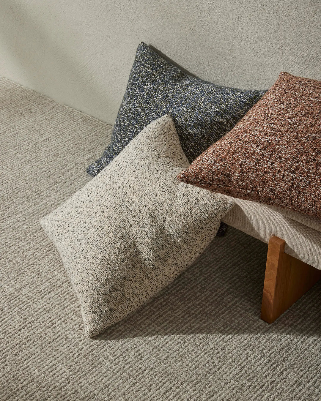 Neutral earth-toned cushion styling with charcoal, oatmeal and terracotta textures on a cream wool rug, mid-century wood frame visible