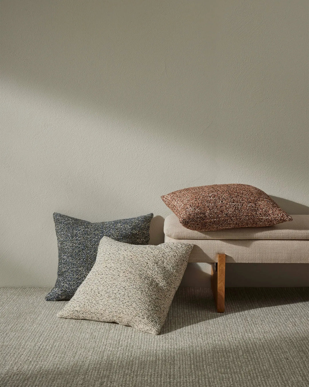 Neutral minimalist scene with Lisette cushions in charcoal, cream and terracotta tones stacked on a wooden-frame bench, set against soft greige walls and natural wool rug