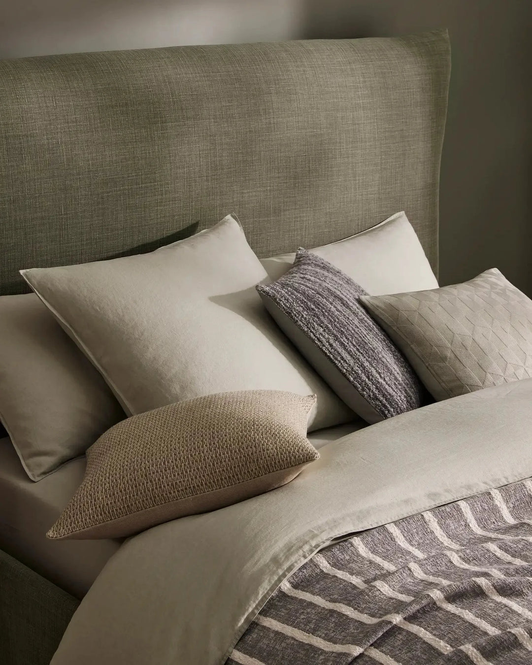 Neutral bedroom scene with layered linen bedding, sage headboard, natural texture cushions in cream and charcoal, and geometric patterned throw in warm grey tones