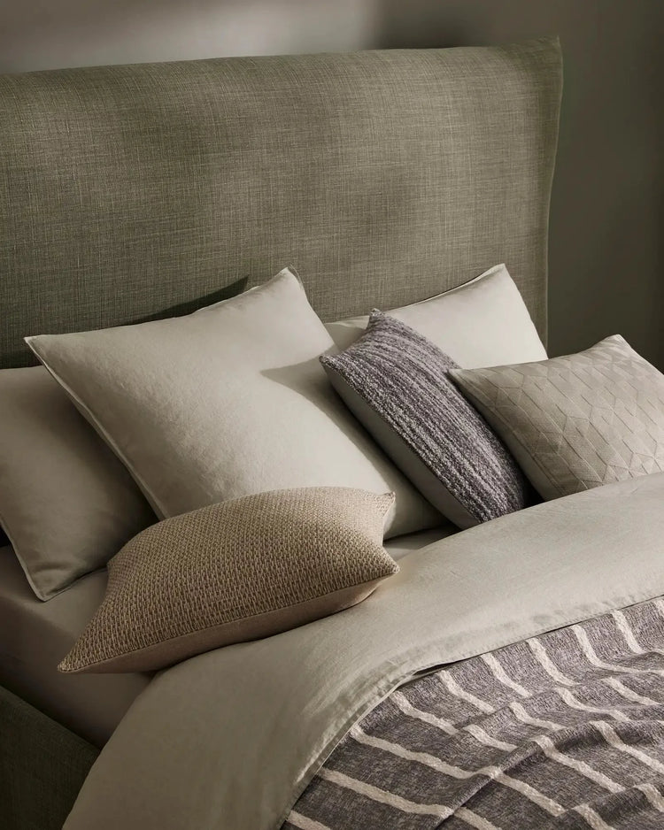 Neutral bedroom scene with layered linen bedding, sage headboard, natural texture cushions in cream and charcoal, and geometric patterned throw in warm grey tones
