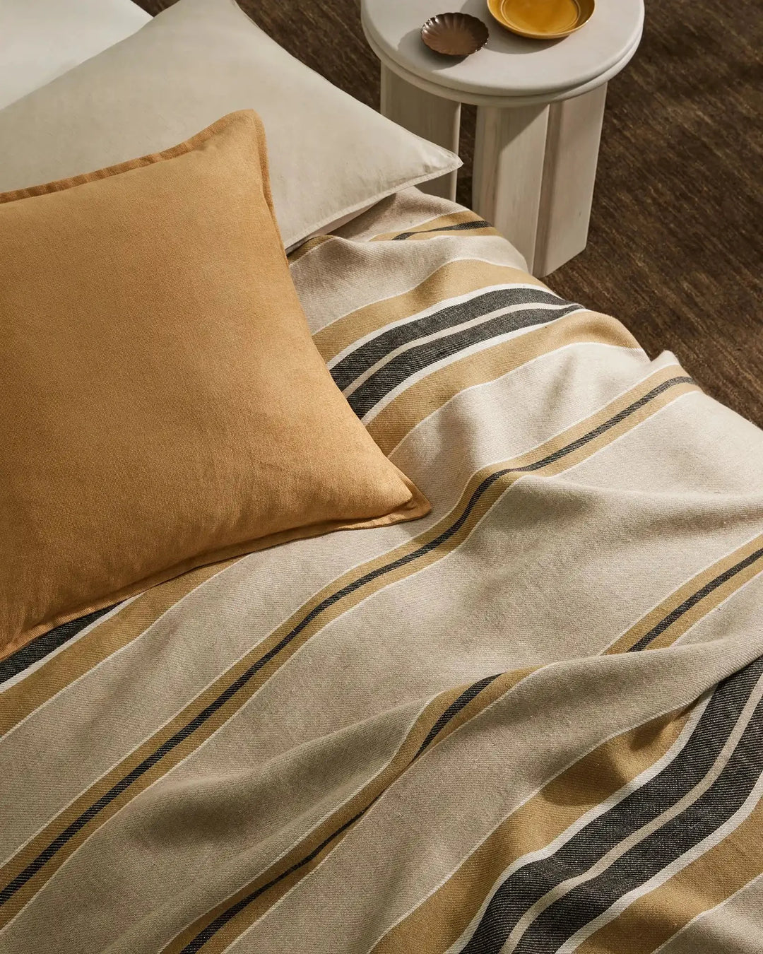 Neutral bedding with ochre and charcoal stripes layered beside a honey-toned linen cushion, styled on a dark wood floor with minimal décor.