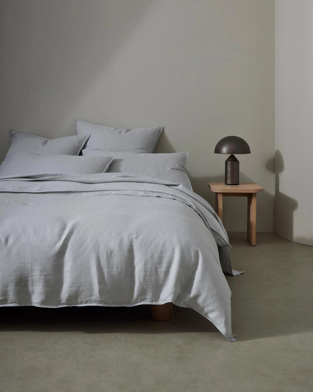 Minimalist bedroom with Ravello linen bedding in soft silver, crisp pillows, and natural wood nightstand with bronze mushroom lamp against neutral walls