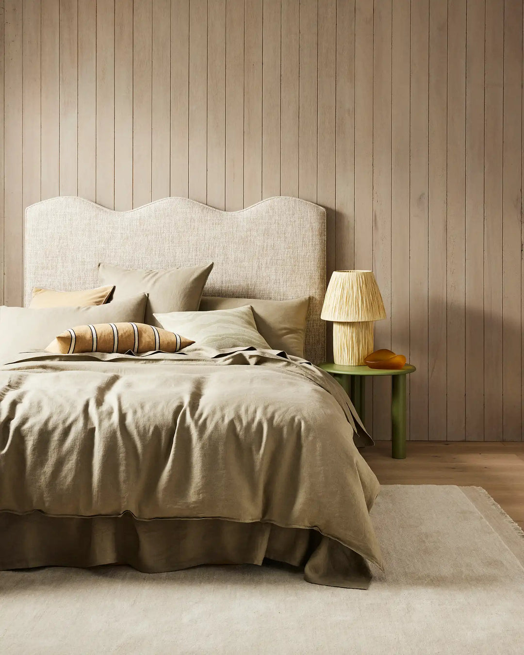 Neutral bedroom with natural linen bedding in cream and sage tones, wavy-edged upholstered headboard, striped linen cushion, and soft area rug creating a warm, minimalist aesthetic