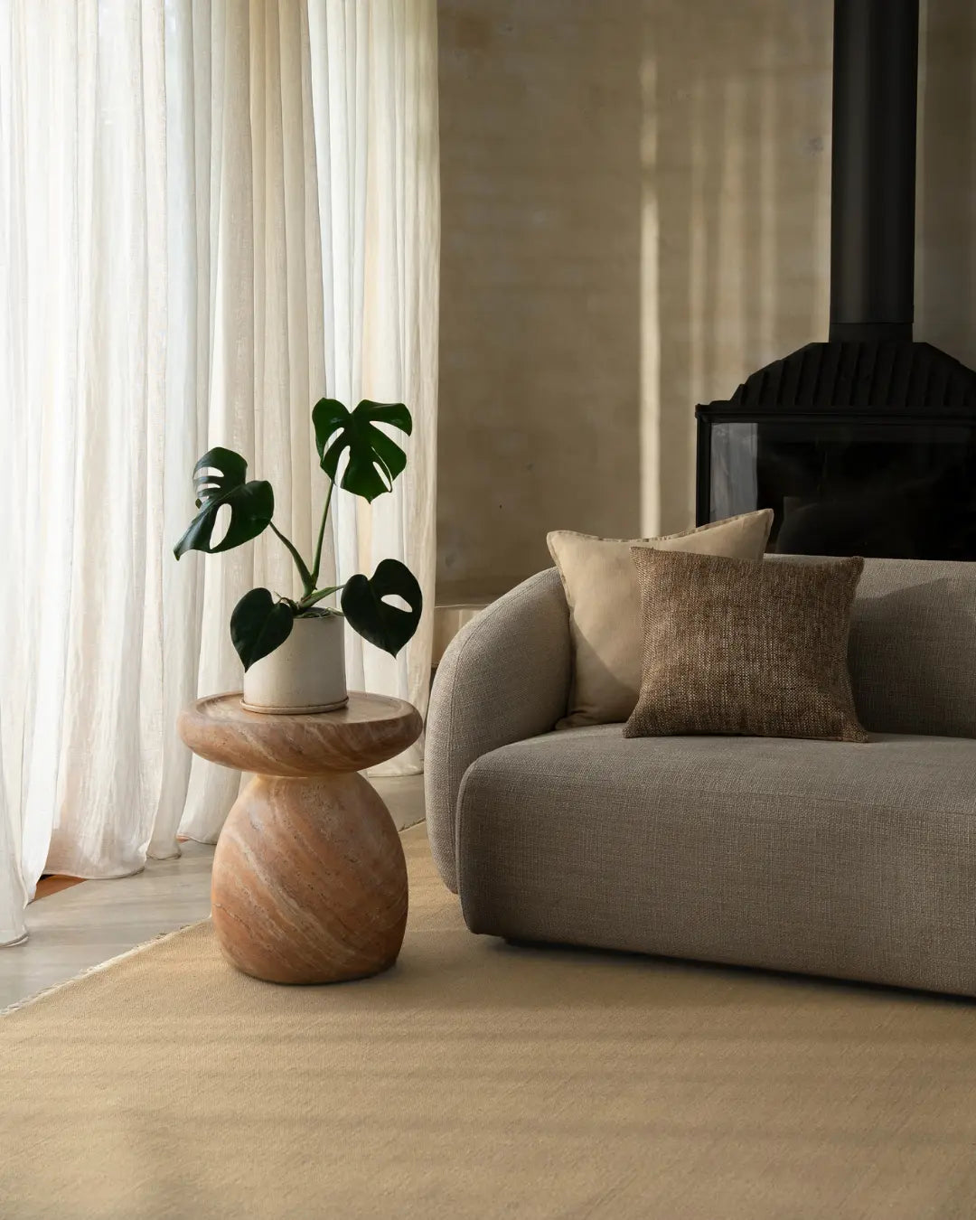 Neutral living room with cream linen curtains, grey upholstered armchair, taupe wool rug, wooden side table, and monstera plant in white pot. Warm minimalist styling with natural light.