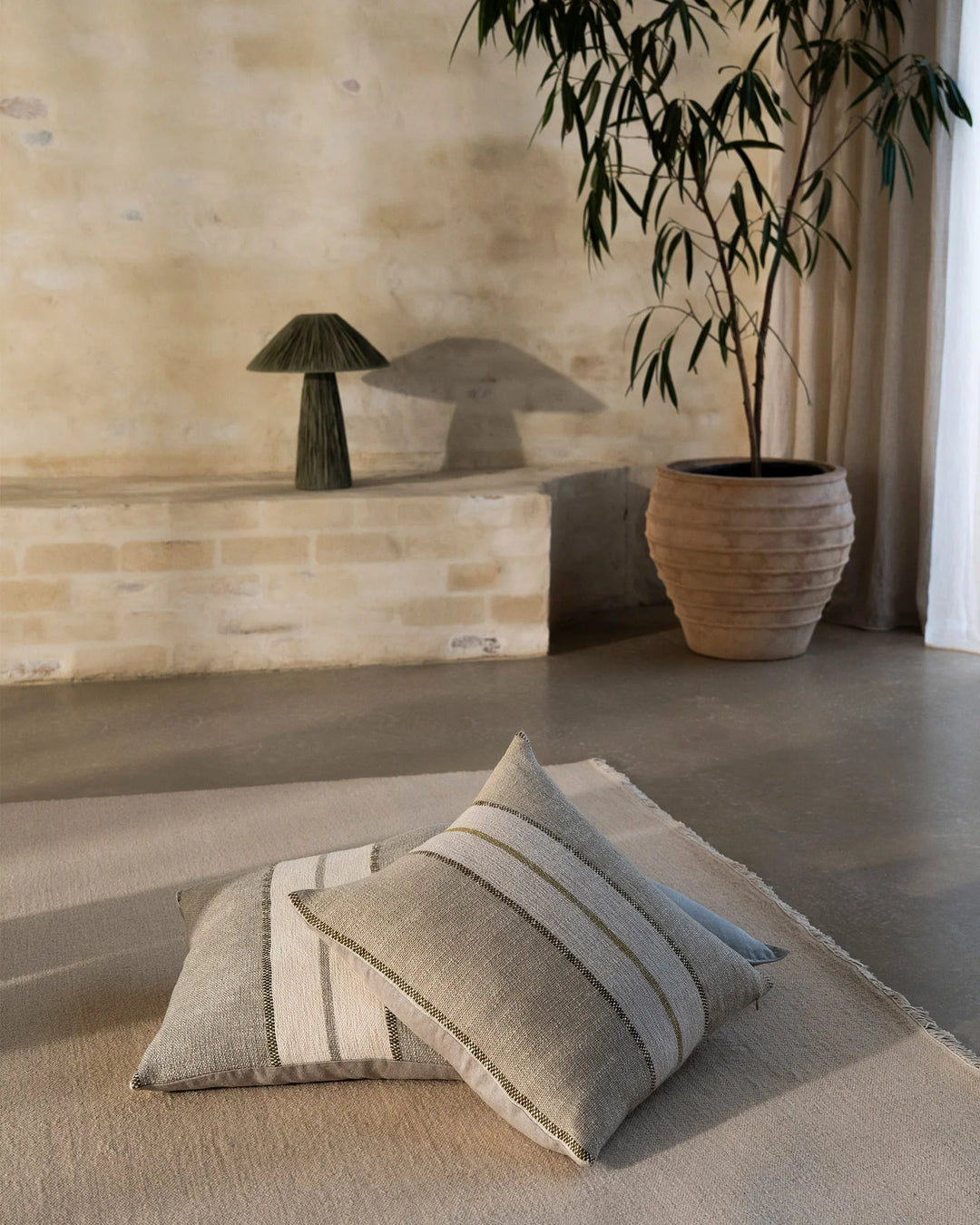Neutral minimalist living space with striped linen cushions on wool rug, featuring sage green potted plant, cream brick wall, and soft natural light