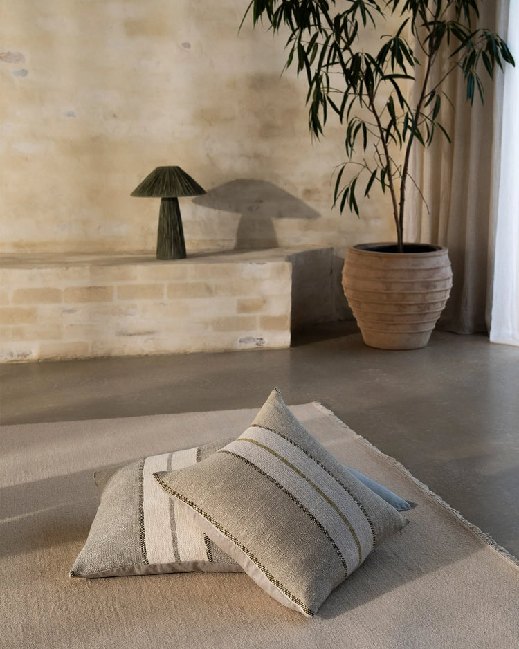 Neutral minimalist living space with striped linen cushions on wool rug, featuring sage green potted plant, cream brick wall, and soft natural light