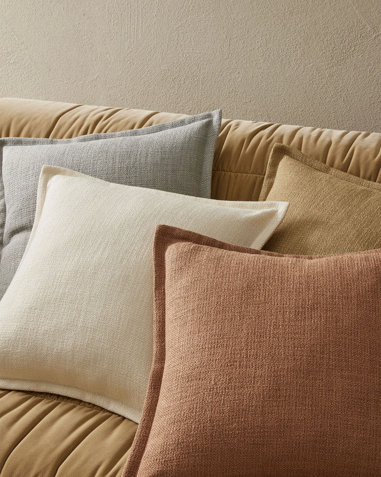 Neutral layered cushion styling on a quilted sofa, featuring cream, grey, terracotta and warm sand textured pillows against a textured beige wall