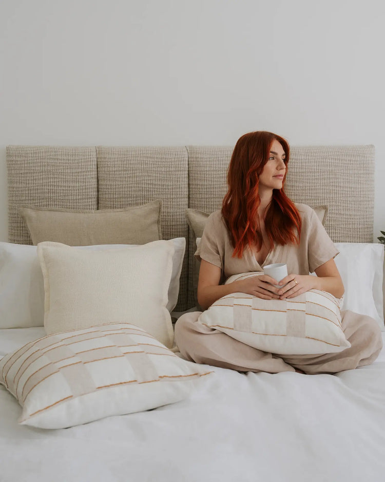 Neutral bedroom scene with woman sitting on bed, featuring textured linen cushions and striped bedding in cream and warm terracotta tones against soft grey upholstered headboard